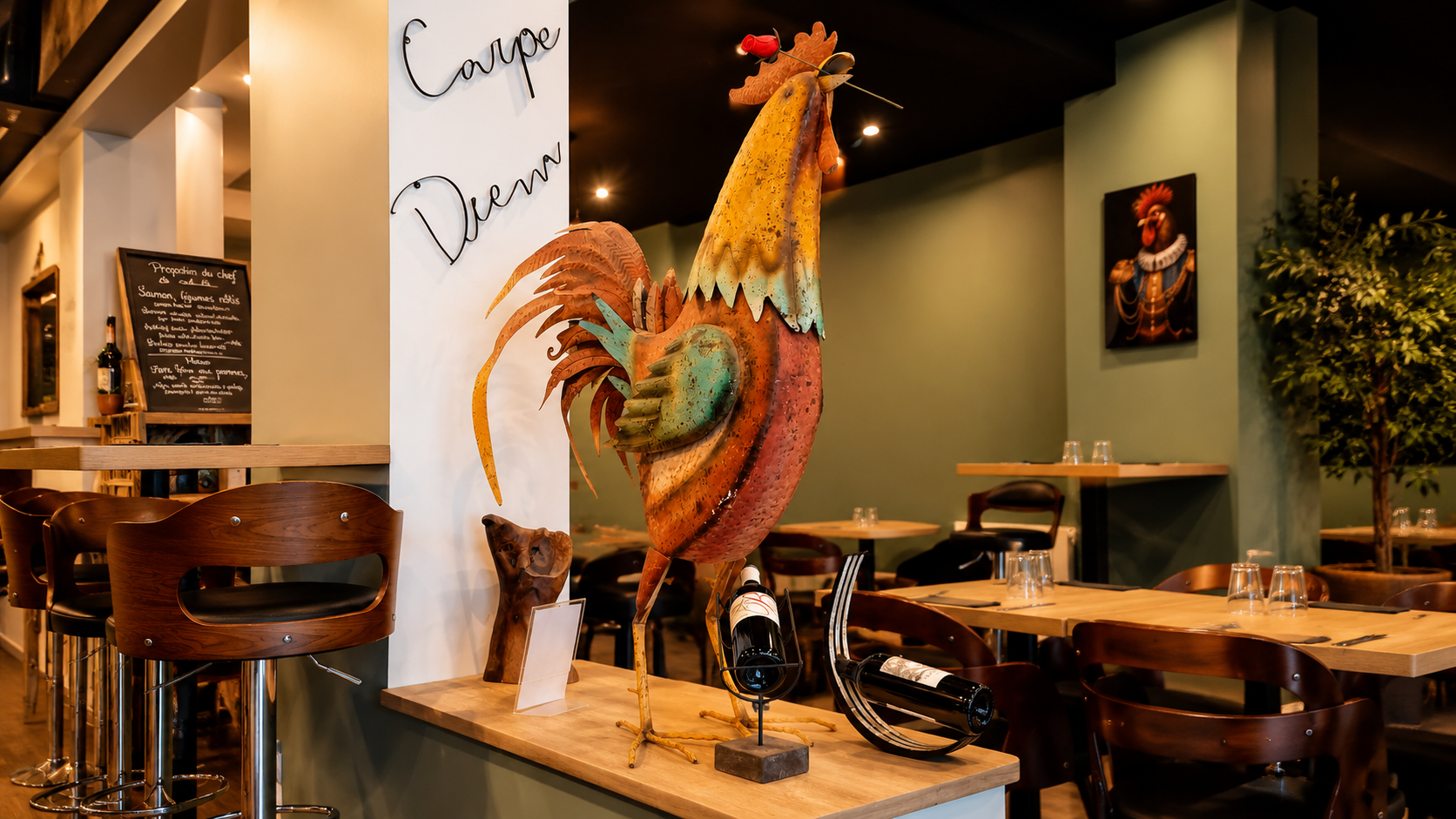 Salle du restaurant O'24 à Dreux : grande sculpture de coq en métal, mur vert végétal, tables en bois et ardoise du chef.